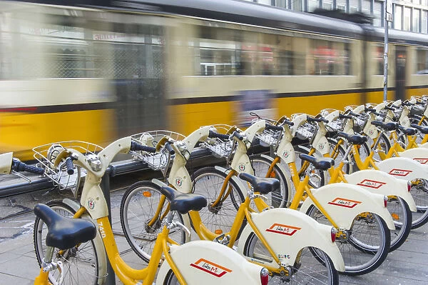 Rows of yellow bike sharing bicycles with a moving tram, Milan, Italy