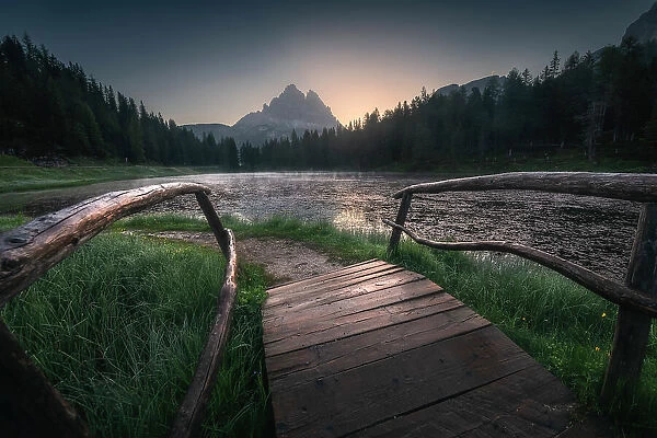 A small wooden bridge leads the eye towards the majestic Tre Cime di Lavaredo and the Antorno lake in the background. Dolomites, Italy