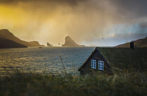 A snow shower is moving towards the turf houses in the village of Bour. In the background Drangarnir and Tindholmur are illuminated by the low sun. Vagar, Faroe Islands