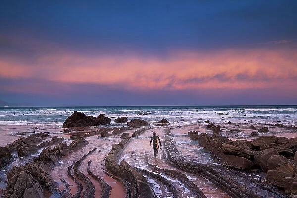 Spain, Basque country, a surfer walking towards the sea at a rock beach near Bilbao at sunrise