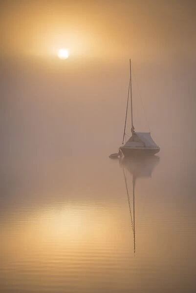 Sun rising over a sailing boat moored on a misty Wimbleball Lake, Exmoor National Park