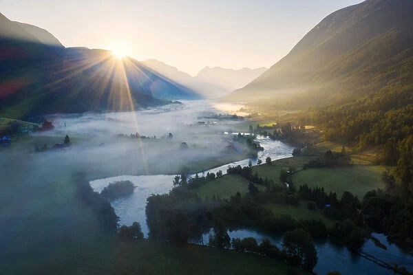 Sunburst during a foggy sunrise over Stryneelva river and fields, aerial view, Stryn