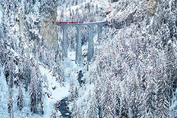 Swiss red train crossing Landwasser viaduct above a frozen river in the snowy landscape, Filisur, Graubunden canton, Switzerland