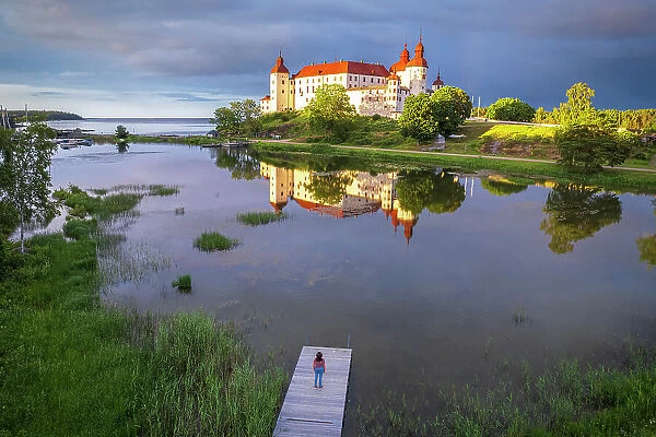 One tourist standing on the river of Vanem lake admires Lacko castle at sunset, Kallandso island, Lidkoping municipality, Gotaland, Sweden