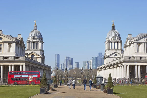 A traditional double-decker red bus passing by the Greenwich University at the Old Royal Naval College with the Canary Wharf skyline in background, London, United Kingdom, Northern Europe