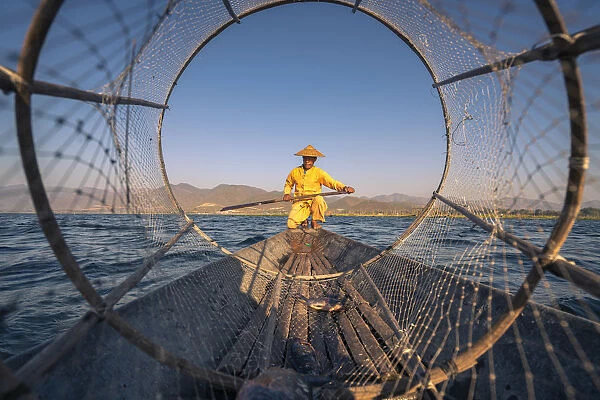 Traditional fisherman viewed through conical fishing net rowing on Lake Inle, Lake Inle
