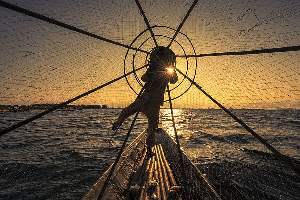 Traditional leg-rowing fisherman viewed through conical fishing net rowing a boat on Lake