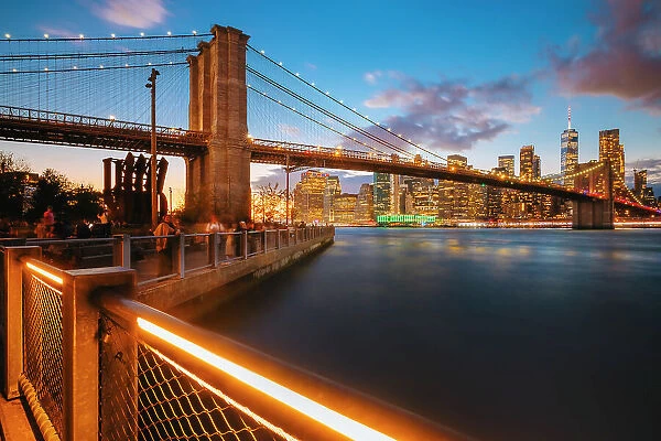 USA, New York City, people enjoying the view of the Brooklyn bridge and the skyscrapers of the financial district illuminated and reflecting in the river