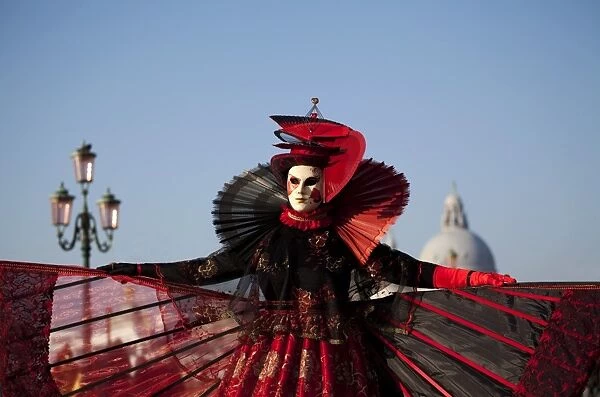 Venice, Veneto, Italy; A mask in costume on the Bacino di San Marco with the cupola of Santa Maria della Salute in