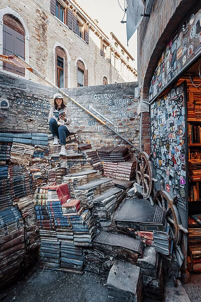 Venice, Veneto, Italy. Woman sitting on books at ancient Acqua Alta Library (MR)