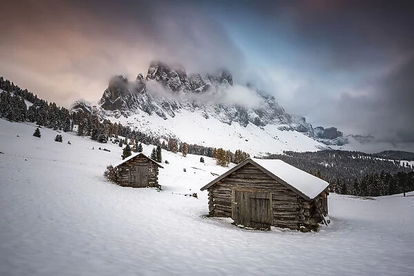 Winter scenery with two wooden lodges and Odle Dolomites covered in snow. Malga Gampen