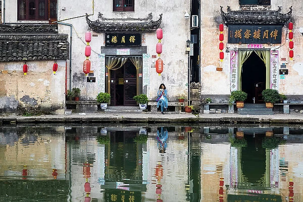 Woman on bench and village buildings reflected in pond, Hongcun, China