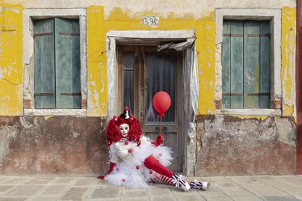 A woman dressed as a clown holds a ballon in front of a colourful facade on Burano