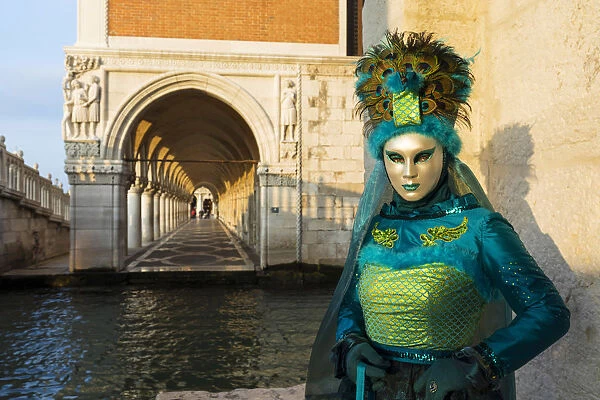 A woman in a mask and green costume stands by the canal next to St Marks square