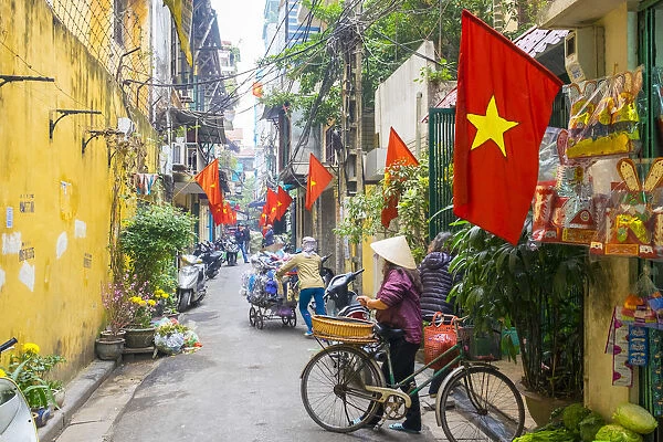 Woman pusing a bicycle through an alleyway lined with red Vietnamese flags, Hoan