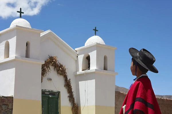 A young Aymara boy wearing traditional clothes stand in front of the small church of