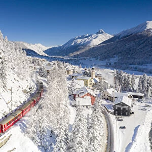 Aerial view of famous red Bernina Express train crossing the snowy village of Madulain