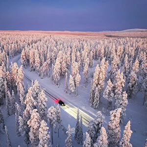 Car traveling on icy road crossing the winter forest covered with snow from above, Lapland, Finland