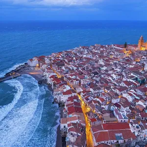 Dusk above the red tile roofs of Cefalu with street lights on and rough sea, aerial view, Cefalu, Palermo province, Tyrrhenian sea, Sicily, Italy Dusk above the red tile roofs of Cefalu with street lights on and rough sea, aerial view, Cefalu, Palermo province, Tyrrhenian sea, Sicily, Italy