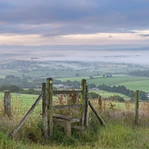 Footpath and stile through farmland with mist covered countryside beyond, Devon, England