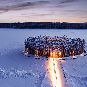 Illuminated circular building of the floating Arctic Bath Hotel and walkway covered with snow at dusk, Harads, Lapland, Sweden
