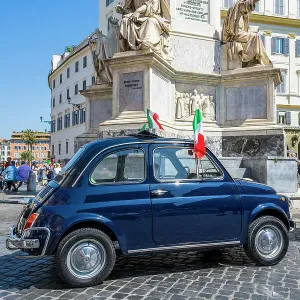 Old iconic blue Fiat 500 car with Italian flags parked in a cobbled square, Rome, Lazio, Italy