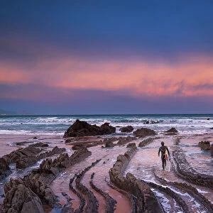 Spain, Basque country, a surfer walking towards the sea at a rock beach near Bilbao at sunrise