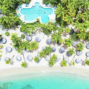 Swimming pool and parasols framed by palm trees on white sand of tropical beach from above, Antigua, Caribbean, West Indies
