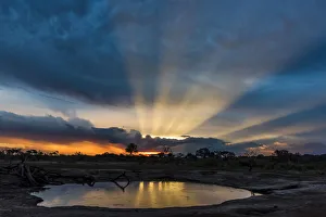 Africa, Zimbabwe, Hwange National park. View across a waterhole at sunbeams bursting