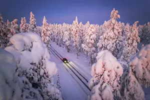 Road Trip Prints: Drone a car with headlight on travelling the icy slippery road crossing the forest covered with snow
