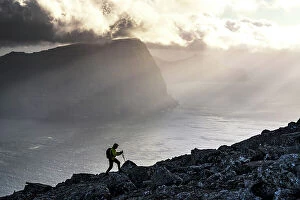 Climate Collection: Man with backpack climbing a mountain above the fjord lit by sun rays at sunset, Vidoy Island