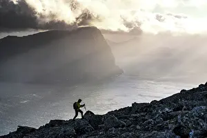 Denmark Prints: Man with backpack climbing a mountain above the fjord lit by sun rays at sunset