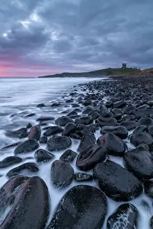 Northumberland Prints: Moody dawn sky over Dunstanburgh Castle from the boulder strewn shore of Embleton Bay