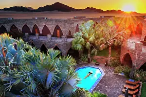 Namibian Prints: Namibia, a woman swimming in a pool in a luxury hotel in Namib Naukluft National Park with the sun setting on the dunes