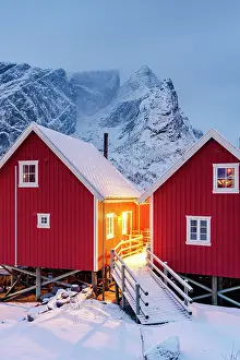 Lofoten Prints: Red wooden cabins at dusk with snowcapped mountains on background