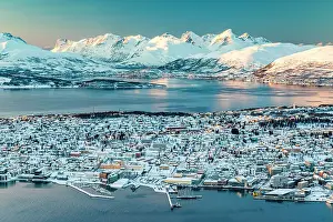 Building Exterior Prints: Snowcapped mountains and icy sea surrounding the frozen harbor and city of Tromso at sunset