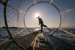 Traditional leg-rowing fisherman viewed through conical fishing net rowing a boat on Lake