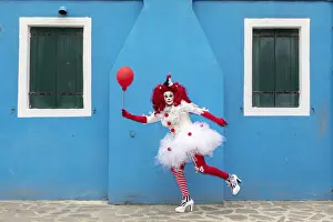 A woman dressed as a clown holds a ballon in front of a colourful facade on Burano