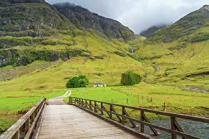 A Charnaich Collection: Wooden bridge leading to cottage under mountains, Glencoe, Scottish Highlands, Scotland, UK